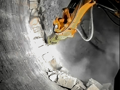 A hydraulic demolition robot with a jackhammer attachment breaks down refractory bricks lining the interior of a large cement kiln.